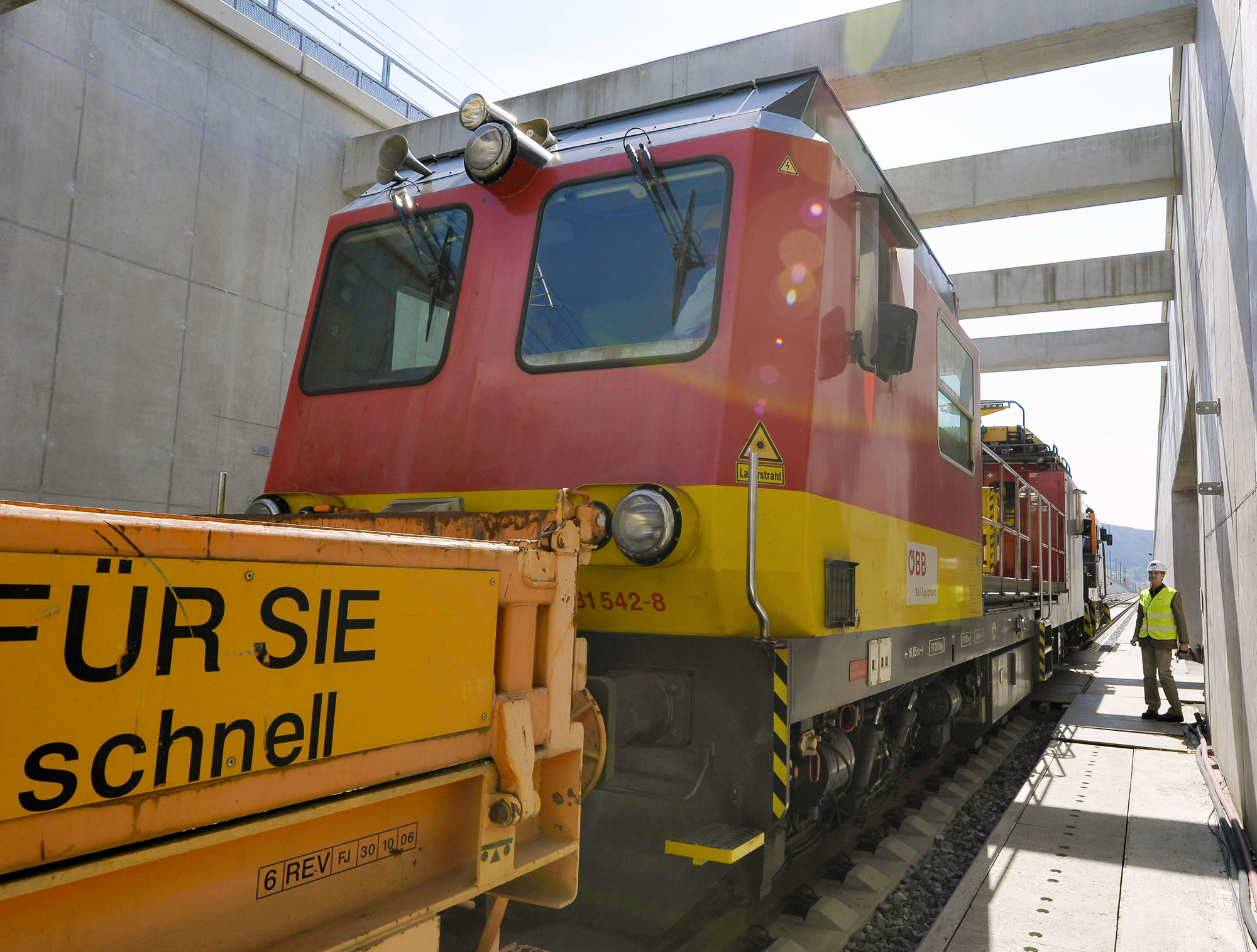 Close-up of a locomotive with a construction worker in safety equipment standing on the right-side platform, overseeing the locomotive.