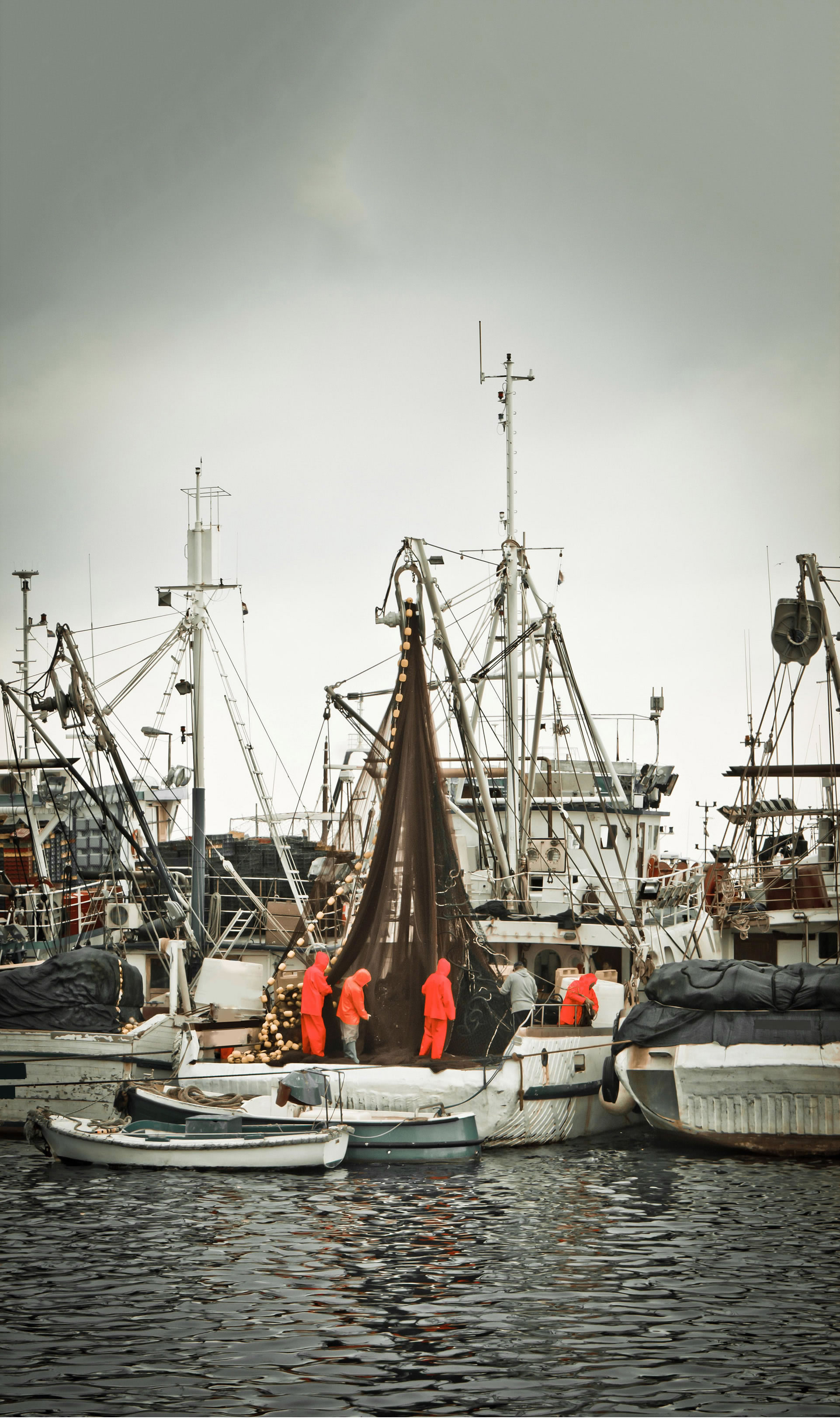 Fishermen in professional suits tending to a fishing net on a trawler, amid a fleet of boats in a fishing harbour.