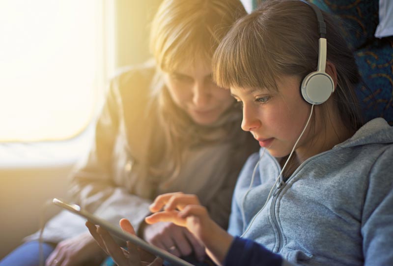 En ung pige med hovedtelefoner leger med sin tablet i toget. Ved siden af hende sidder en kvinde.