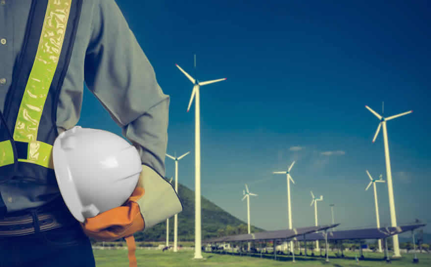 En ingenjör klädd i en overall med reflexdetaljer står och håller i sin skyddshjälm. I bakgrunden ser man ett vindkraftverk. © Fotolia