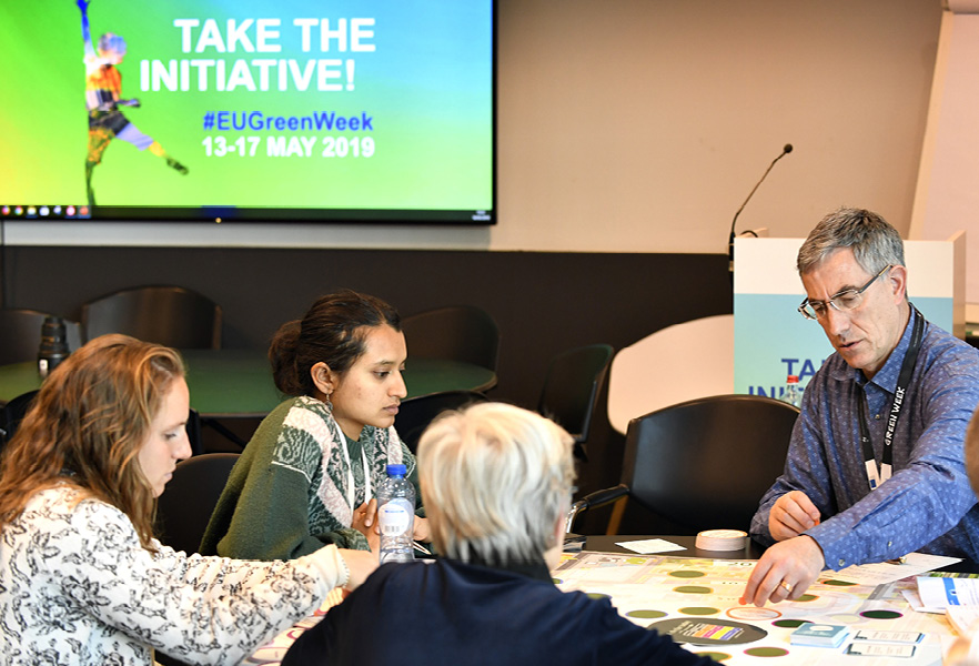 Keskustelijoita tutkimassa esitteitä pöydän ääressä taustallaan näyttö, jolla esitellään vihreästä viikosta kertovaa tietoa