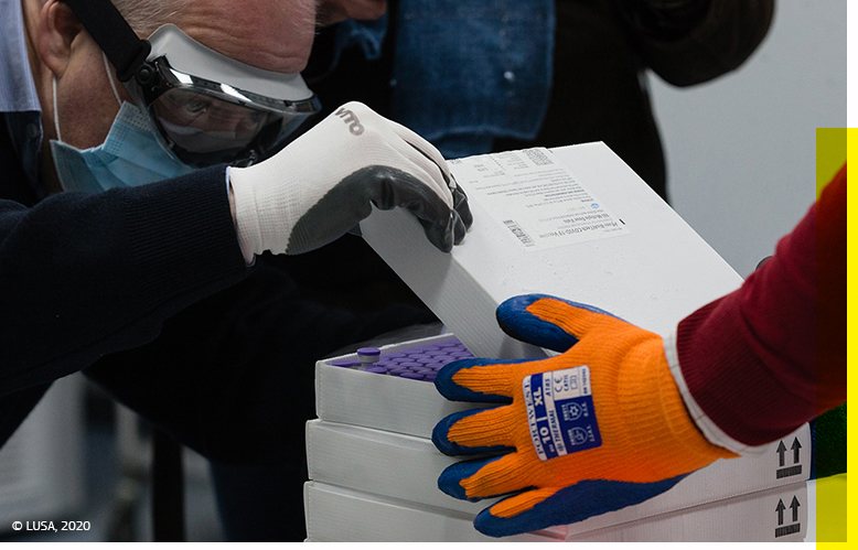 Närbild på en man i skyddsutrustning som öppnar den översta lådan i en stapel med lådor som innehåller injektionsflaskor, samtidigt som han övervakas av en kollega i skyddsutrustning. © Lusa 2020