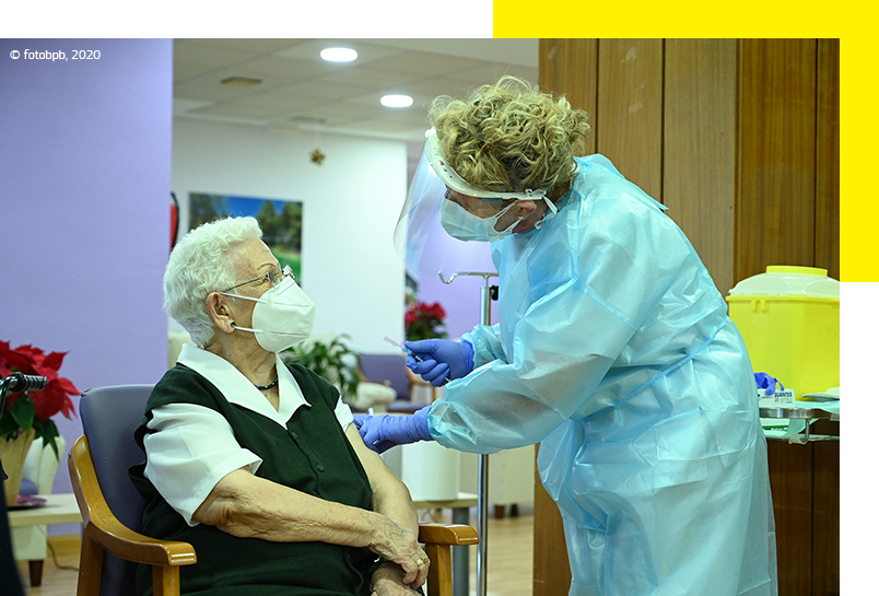 Une femme âgée portant un masque reçoit un vaccin administré par une travailleuse du secteur de la santé équipée d’une tenue de protection complète. © fotobpb, 2020