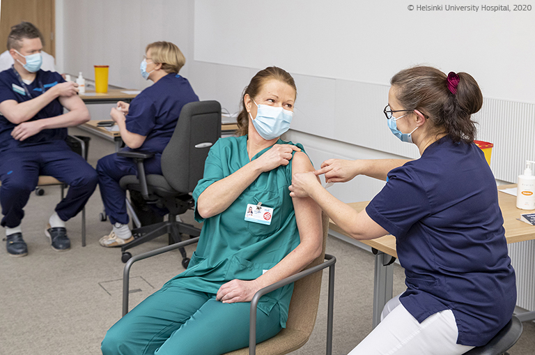 Kolegė skiepija medicinos darbuotoją su darbo drabužiais ir kauke, antrame plane skiepijami kiti medicinos darbuotojai. © Helsinkio universiteto ligoninė, 2020.