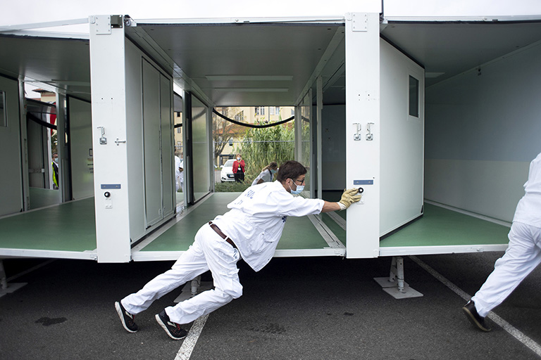 Un bărbat în uniformă medicală albă împinge printr-o parcare săli de consultație modulare temporare.