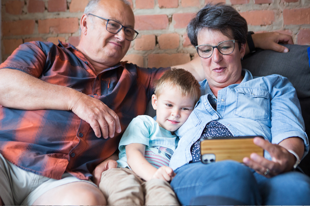 Dois adultos e uma criança pequena estão sentados num sofá e olham para um telemóvel.