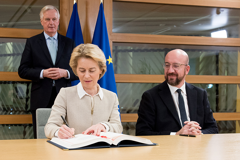 Ursula von der Leyen e Charles Michel assinam o acordo, vendo-se Michel Barnier de pé, ao fundo.